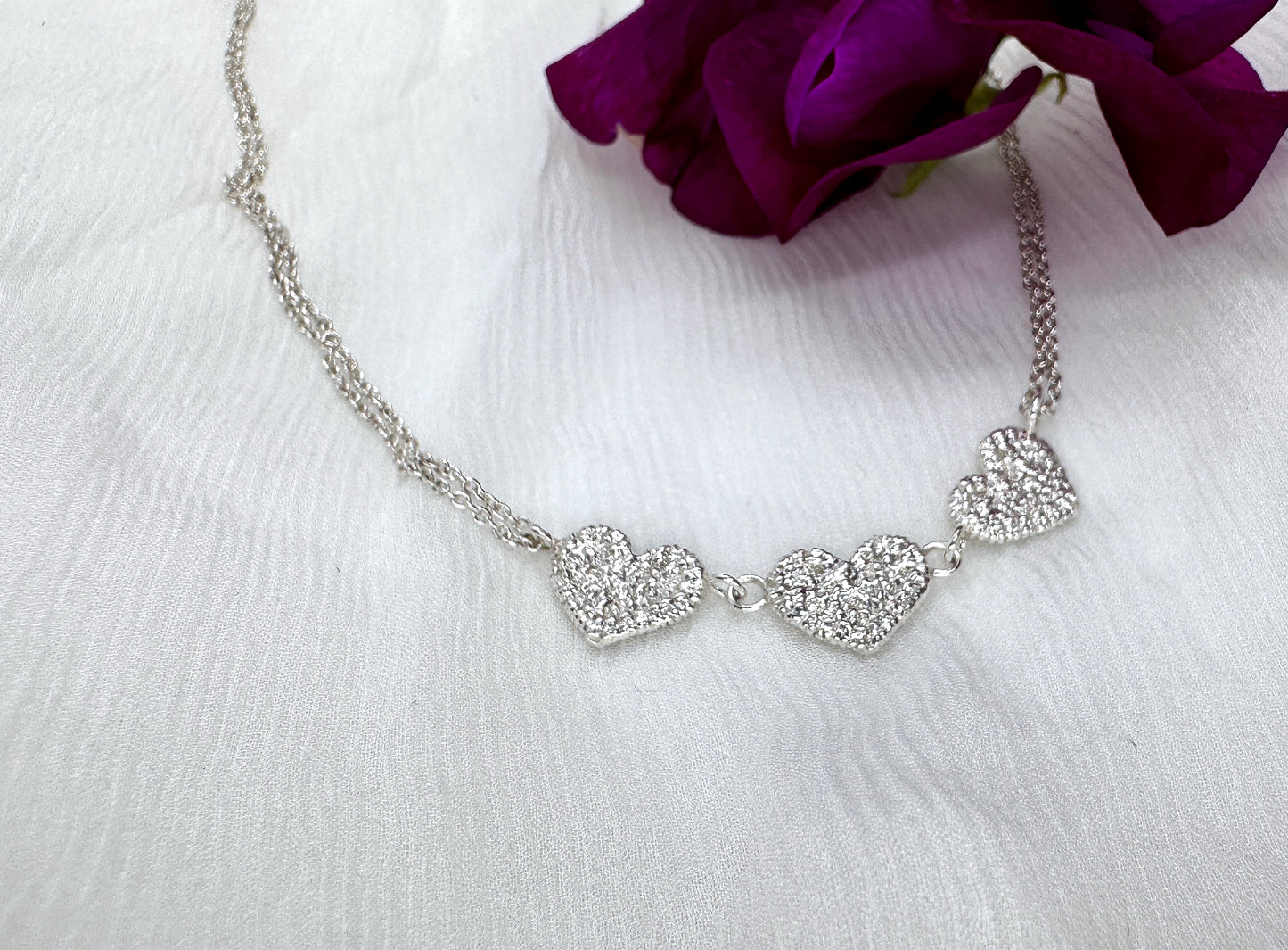 A silver necklace sits on top of a white muslin. The necklace has three textured hearts in a row, originally stitched in lace and then cast in silver. It is on a two-strand chain. On the top right of an image, there is a dark purple flower.