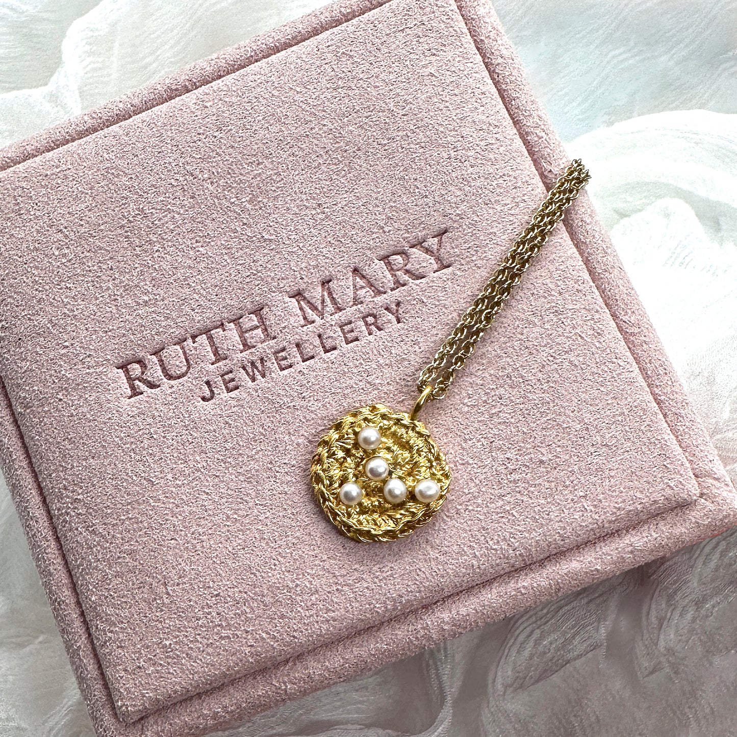 A gold necklace laid across a pale pink jewellery box from Ruth Mary Jewellery. The pendant is a textured circular twist, originally stitched in lace and then cast in gold. It is set with a cluster of small round pearls.