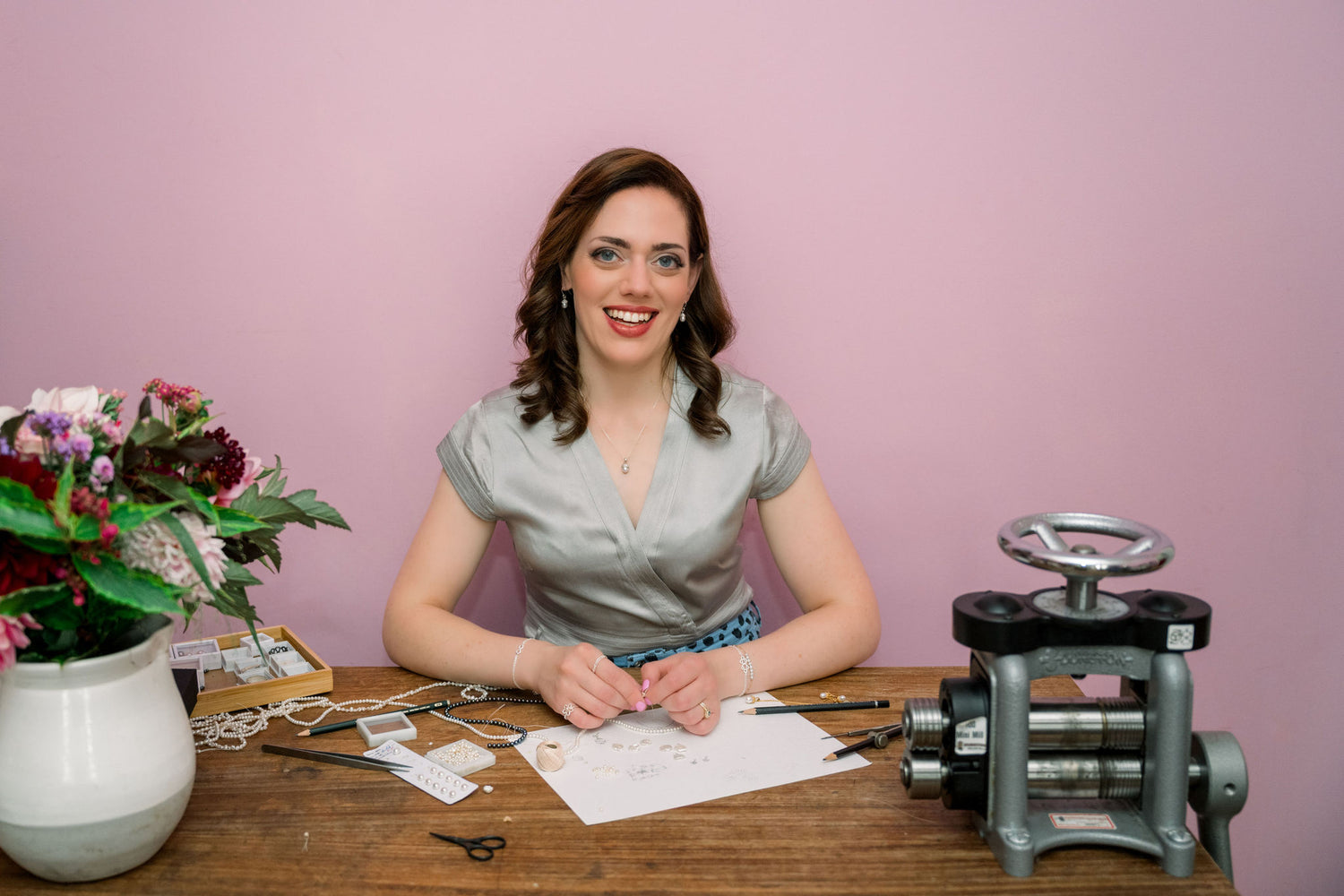 Smiling woman sitting at a table with floral arrangements and jeweller's tools against a pink background. In front of her are a selection of pearls. She is wearing jewellery from her own collection.
