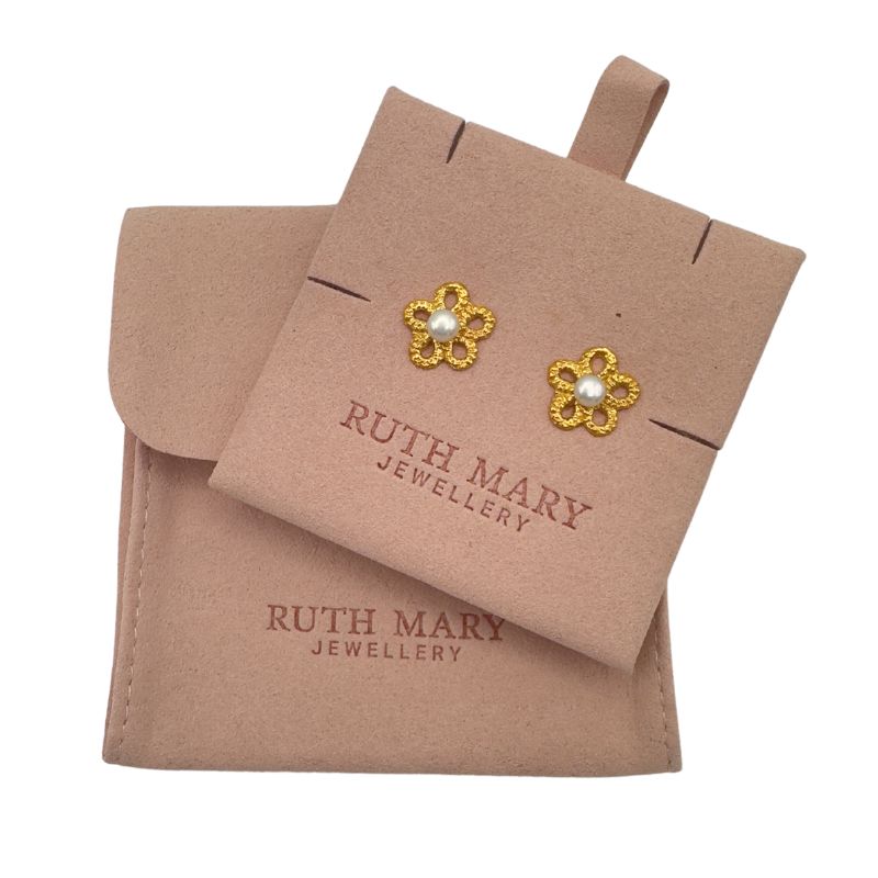 A pale pink textured jewellery pouch. Attached to the pouch are two gold, flower shaped earrings. The centre of the earrings are set with pearls.
