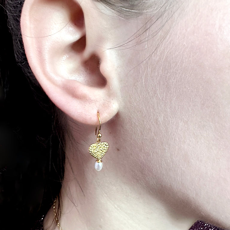 The side of a woman's face. The focus of the image is the earring in her ear. The earring motif is a textured gold heart, originally stitched in lace and then cast in gold. Under the heart hangs an oval-shaped pearl.
