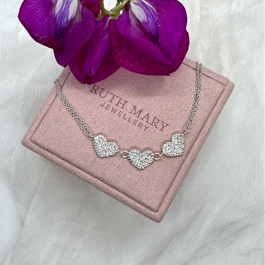 A silver necklace sits on top of a pale pink jewellery box from Ruth Mary Jewellery. The necklace has three textured hearts in a row, originally stitched in lace and then cast in silver. It is on a two-strand chain.