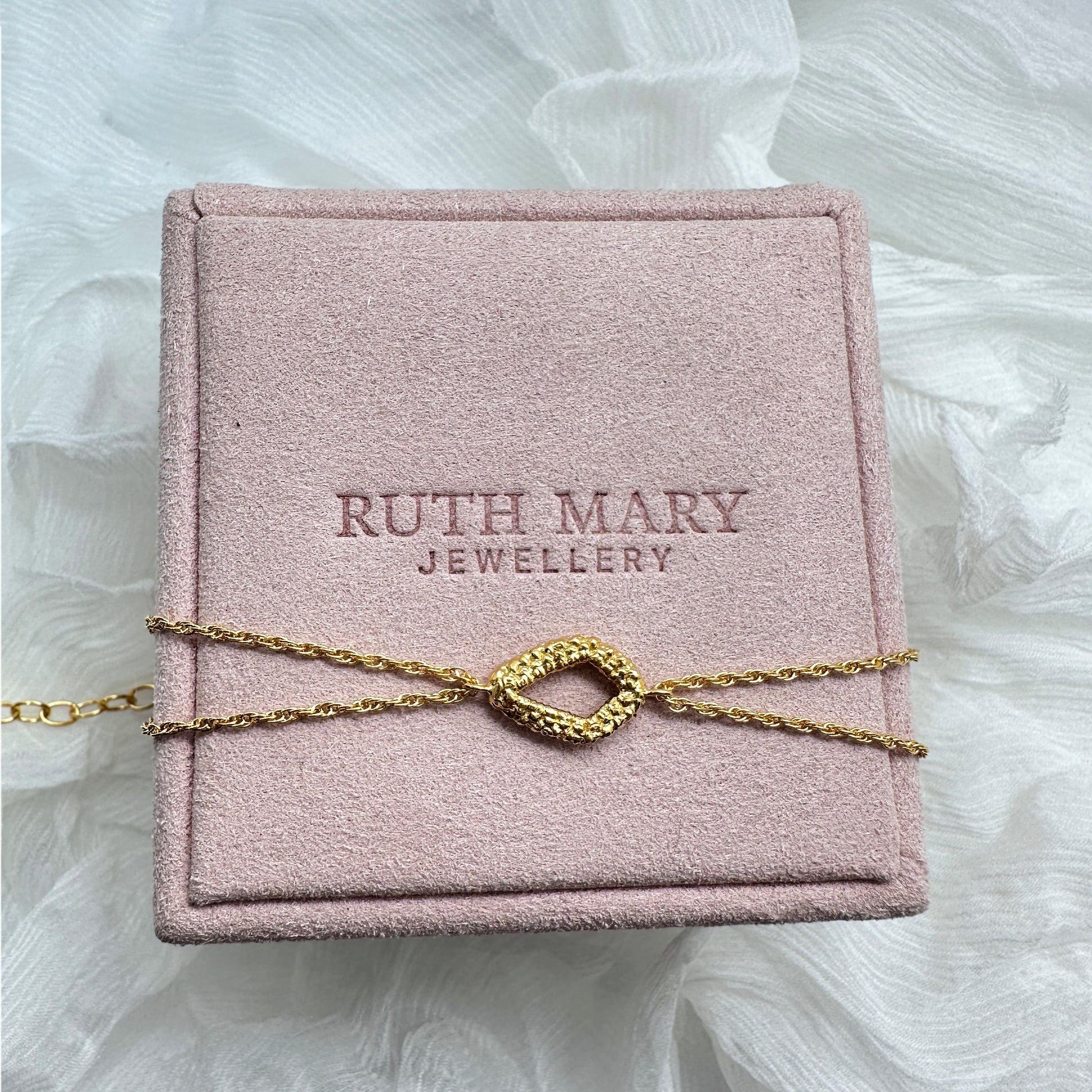 A bracelet laid on top of a pale pink jewellery box from Ruth Mary Jewellery. The motif is a diamond shape, originally stitched out of lace and then cast in gold. The bracelet chain has two strands.