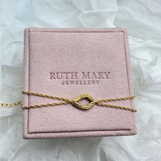 A bracelet laid on top of a pale pink jewellery box from Ruth Mary Jewellery. The motif is a diamond shape, originally stitched out of lace and then cast in gold. The bracelet chain has two strands.
