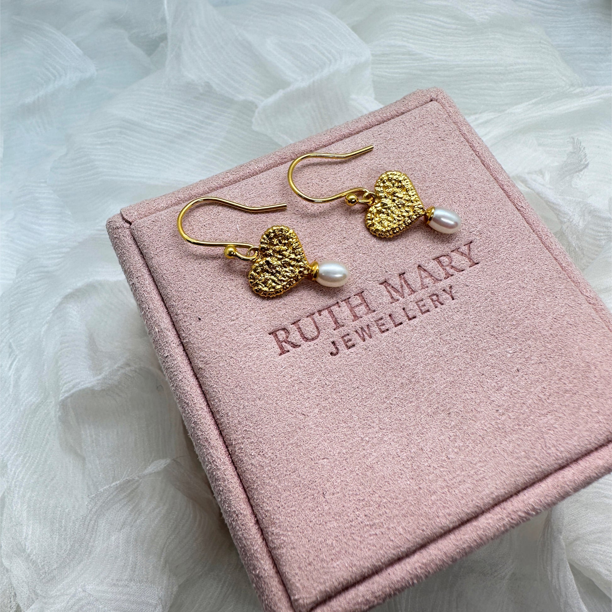 On a pale pink jewellery box there is a pair of earrings. The earring motif is a textured gold heart, originally stitched in lace and then cast in gold. Under each heart hangs an oval-shaped pearl.