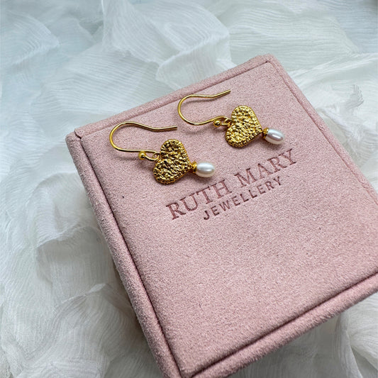 On a pale pink jewellery box there is a pair of earrings. The earring motif is a textured gold heart, originally stitched in lace and then cast in gold. Under each heart hangs an oval-shaped pearl.