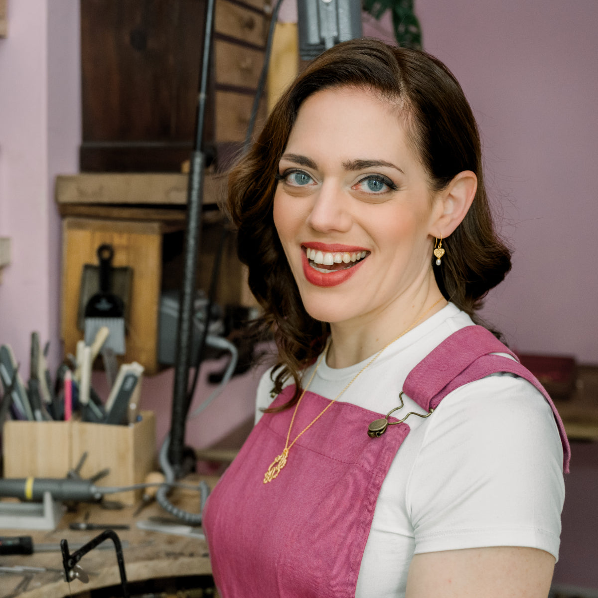 A woman with large bule eyes and dark, curly hair, smiles at the camera. She's wearing raspberry-coloured cord dungarees and gold heart and pearl earrings. She has an unusual necklace on.
