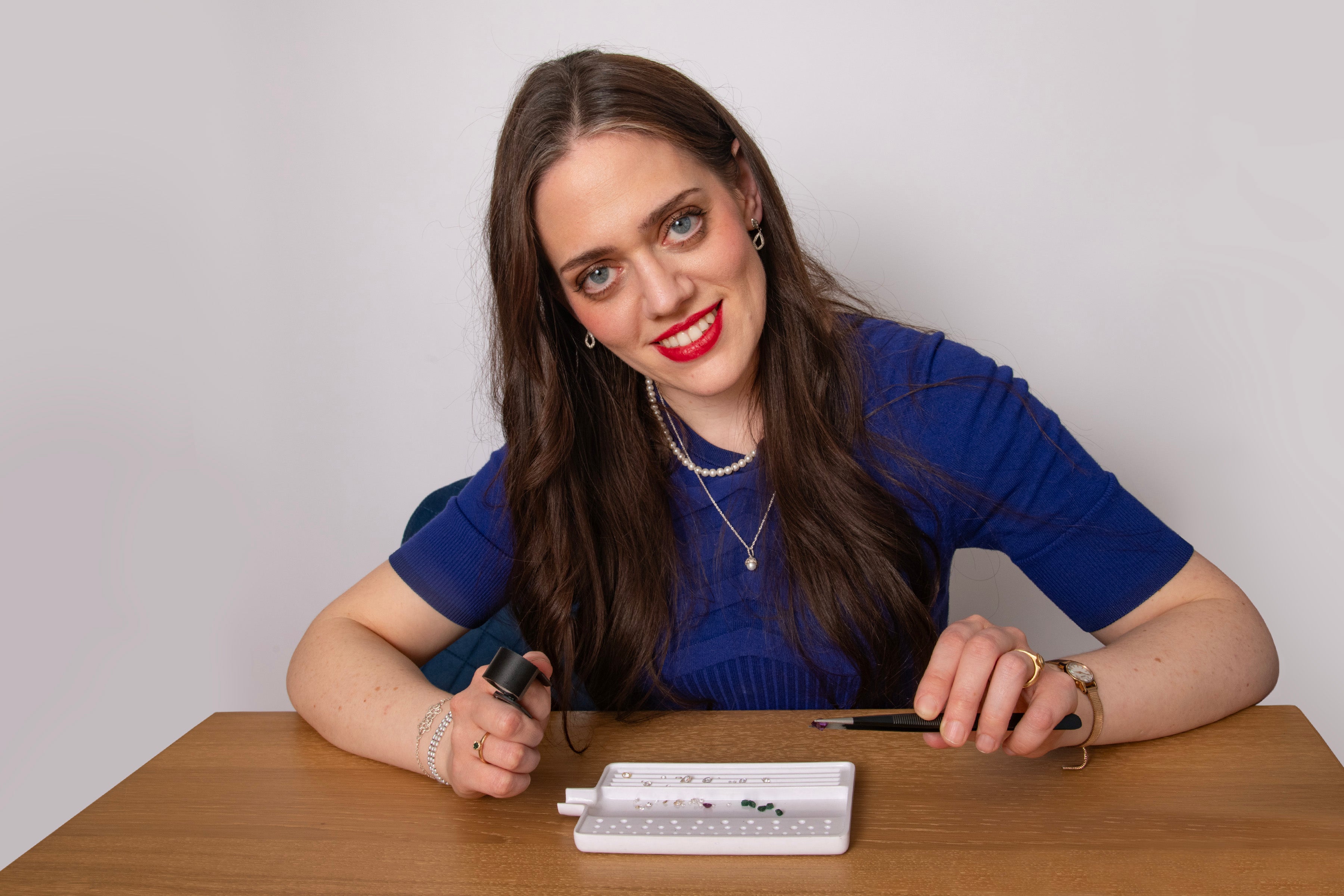A young woman looks directly at the camera, smiling. She has large blue eyes and long brown hair. She is wearing red lipstick and a dark blue top. She is holding loupe and a pair of tweezers, ready to examine a precious gem. In front of her on a wooden table is an open tray filled with various gems. 