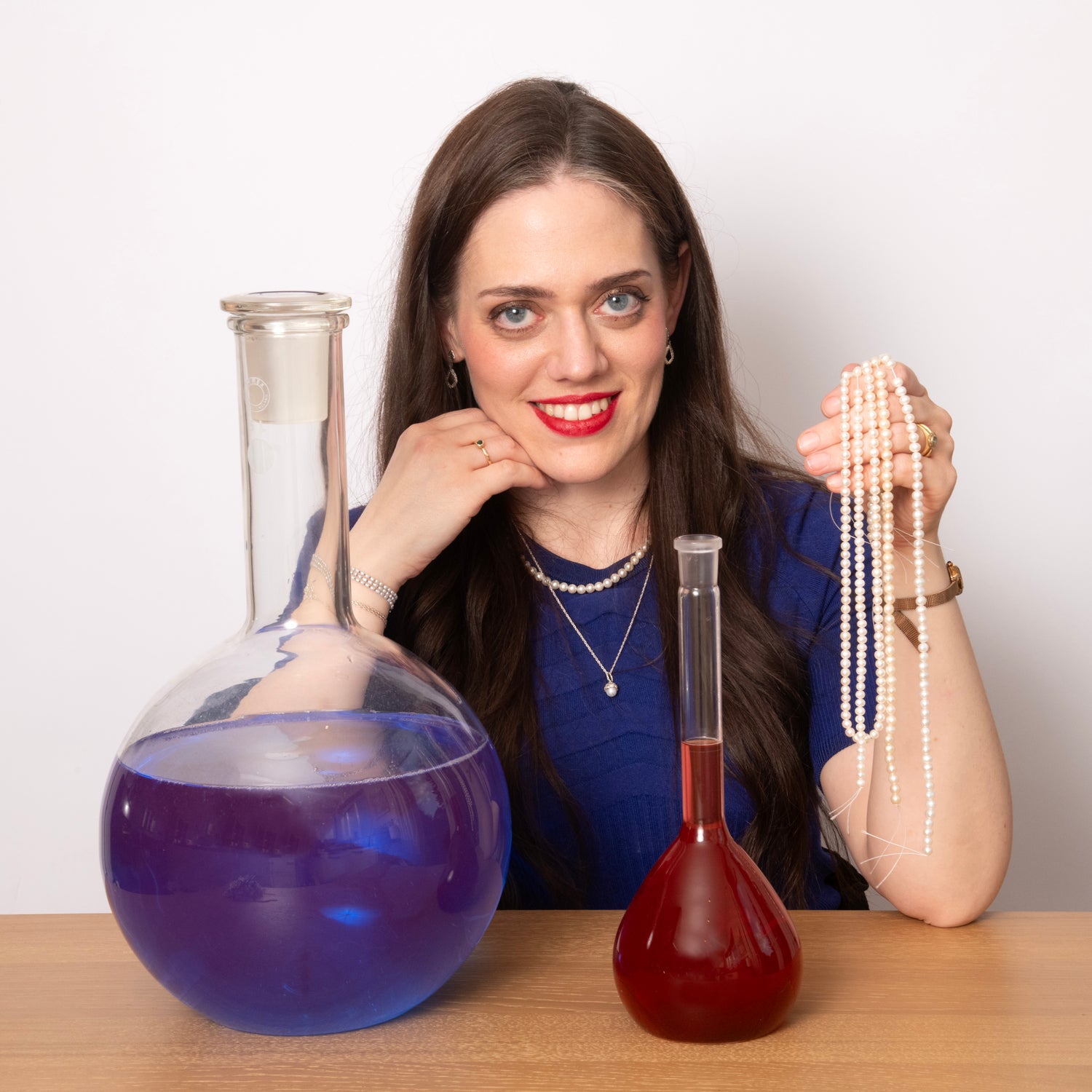 A young woman with long, dark hair sits behind a wooden table. She has blue eyes and a dark blue top on. She is looking directly at the camera. In her right hand, she is holding a string of pearls. In front of her are two chemistry flasks, one filled with purple liquid and the other filled with red liquid.