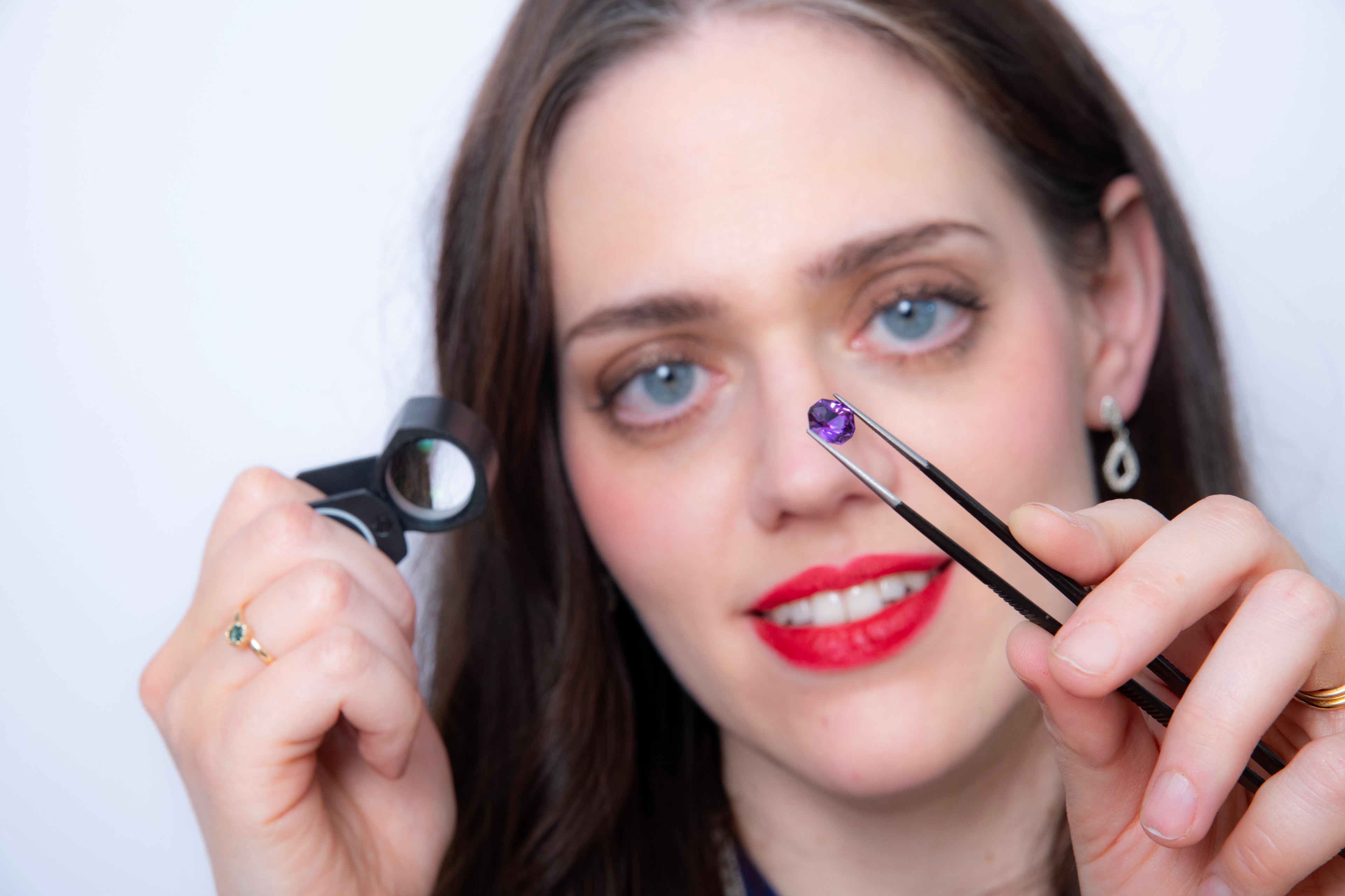 A woman with dark hair and blue eyes is examining a bright purple gem. She is holding the jewel in tweezers and using a jeweller's loupe to look closely at the gem.