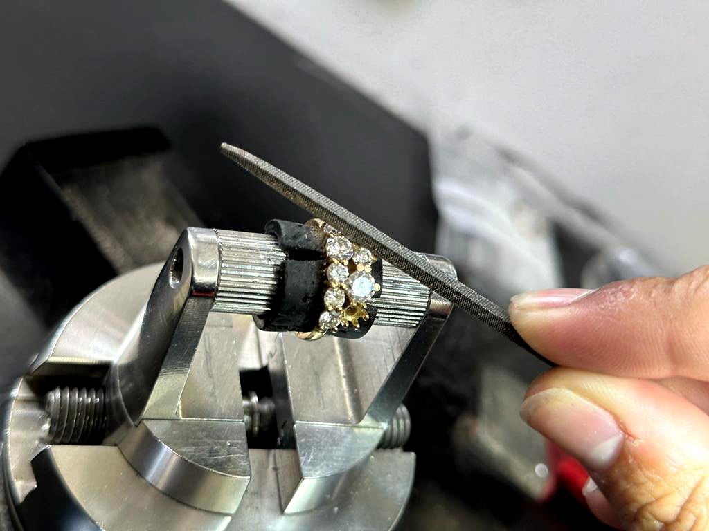 A diamond rings is on a jeweller's tool. We can see the jeweller's fingers using a file to repair the ring. A few diamonds are missing.