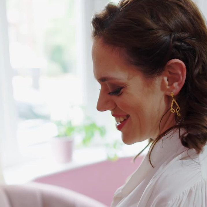 A woman with dark curly hair smiles as she adjusts her unusual geometric earrings. She's wearing a white blouse.